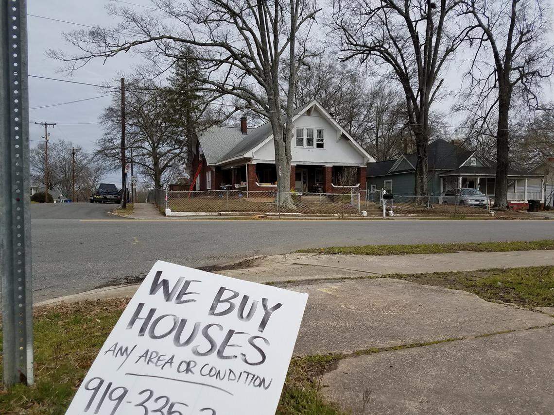 A sign posted at a street corner on Driver Street offers to buy homes. A house across the street is under renovation.