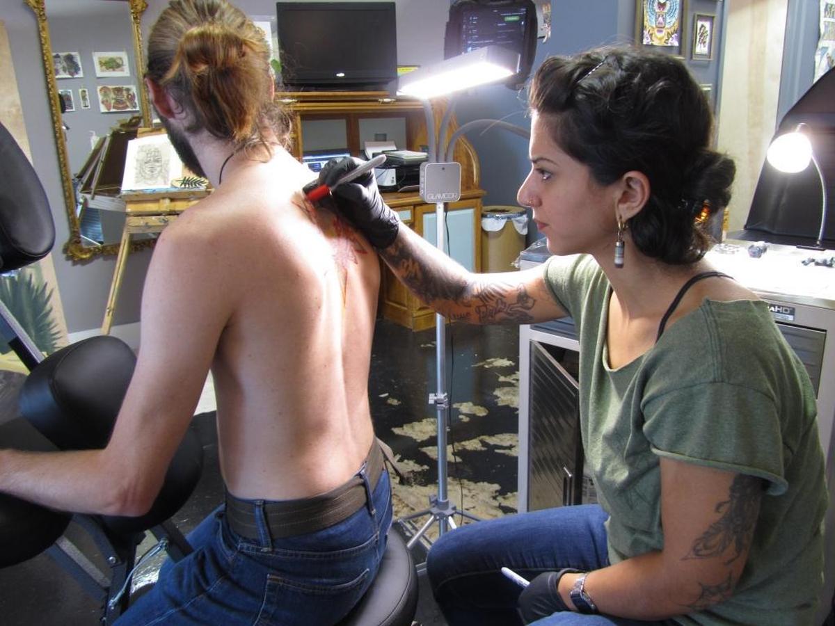 Samantha Castrovinci enhances a tattoo for free on customer Soren Johnsen’ back to add subtle colors to a circle of spear points and lizard design she completed several years ago after only six months’ of experience.