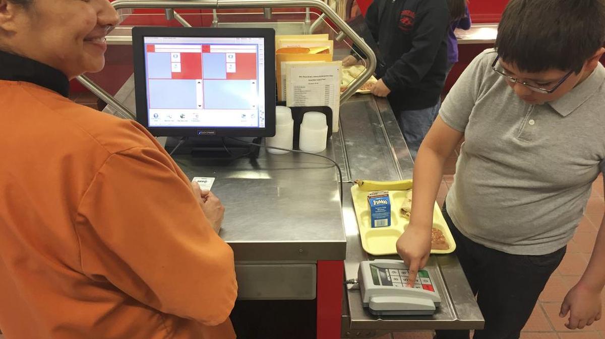 In this Thursday, May 4, 2017 photo, cafeteria worker Paula Herrera, left, supervises as second grade student Dylan Casias punches in his student identification number to deduct a meal payment at Gonzales Community School in Santa Fe, N.M. All students are offered the same lunch at Gonzales and other Santa Fe public schools to avoid any chance of embarrassing students whose parents may have fallen behind on meal payments. New Mexico in April became the first state to outlaw the shaming of children for any unpaid meals.